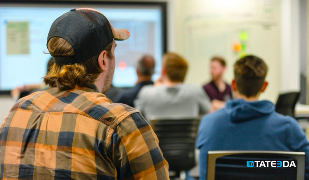 A group of software QA testers attentively listens to their team leader in a small conference hall. The focus is on the testers as the team leader draws a plan on a whiteboard, which is slightly blurred in the background. | TATEEDA