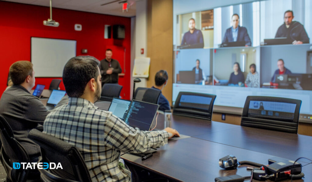 IT professionals in a conference room engage in a video call with remote colleagues, displayed on a large screen, in a modern office setting with vibrant red walls. | TATEEDA