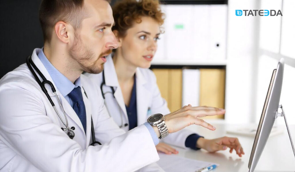 Two medical professionals in white coats discussing healthcare IT solutions on a desktop computer. Highlights how to choose a healthcare IT consulting company to improve workflows and implement innovative systems. | TATEEDA